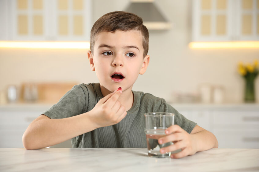 Little boy with glass of water taking vitamin capsule in kitchen