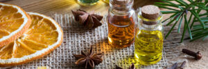 election of essential oils with star anise, clove, cinnamon sticks, frankincense resin, dried orange slices and fir and pine branches in the background