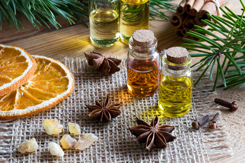election of essential oils with star anise, clove, cinnamon sticks, frankincense resin, dried orange slices and fir and pine branches in the background