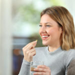 Happy blonde girl taking a vitamin pill and looking at side sitting on a couch in the living room at home