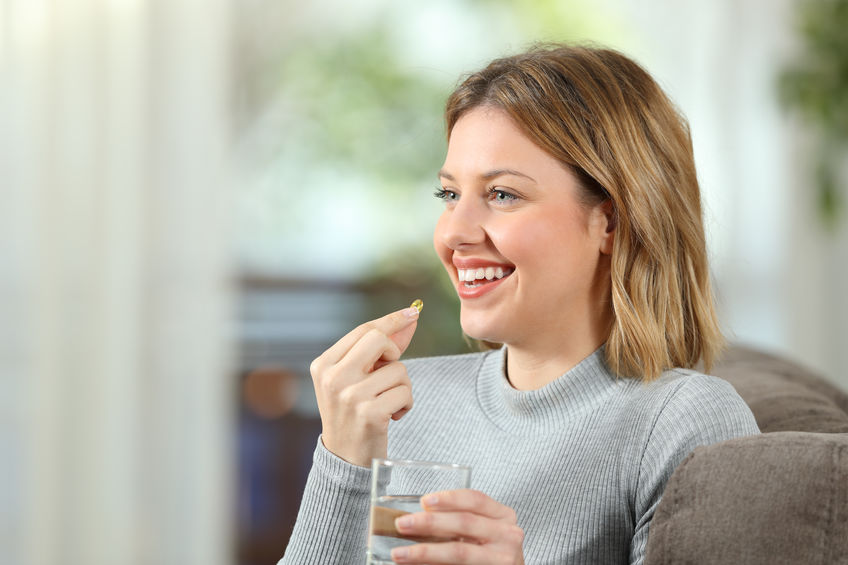 Happy blonde girl taking a vitamin pill and looking at side sitting on a couch in the living room at home