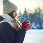 Young Girl in Red Gloves holds a Red Cup with Hot Tea on the Winter Landscape Background with Sunset