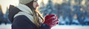 Young Girl in Red Gloves holds a Red Cup with Hot Tea on the Winter Landscape Background with Sunset