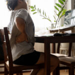 Working woman sitting on the chair in the living room at home She used both hands to press down on the lower back. She is suffering from back pain from sitting for a long time.