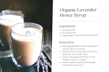 Two glasses of latte on a table next to a recipe card for Organic Lavender Honey Syrup with listed ingredients and directions.
